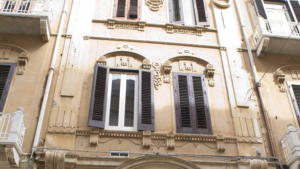 Un edificio con puerta negra, varias ventanas con contraventanas y detalles arquitectónicos decorativos.