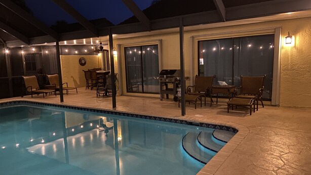 Ein Pool mit überdachtem Patio-Bereich, Gartenmöbeln und einem Grill.