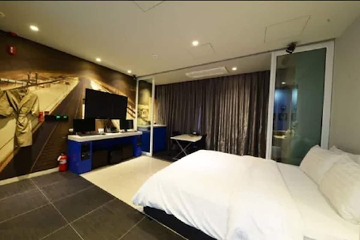 A hotel room with a bed, a desk with a computer, a flat-screen TV, and a large window with curtains.
