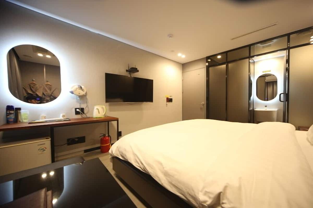A modern hotel room with a large bed, a desk with a flat-screen TV, a mirror, a wardrobe, and a bathroom with a toilet and sink.
