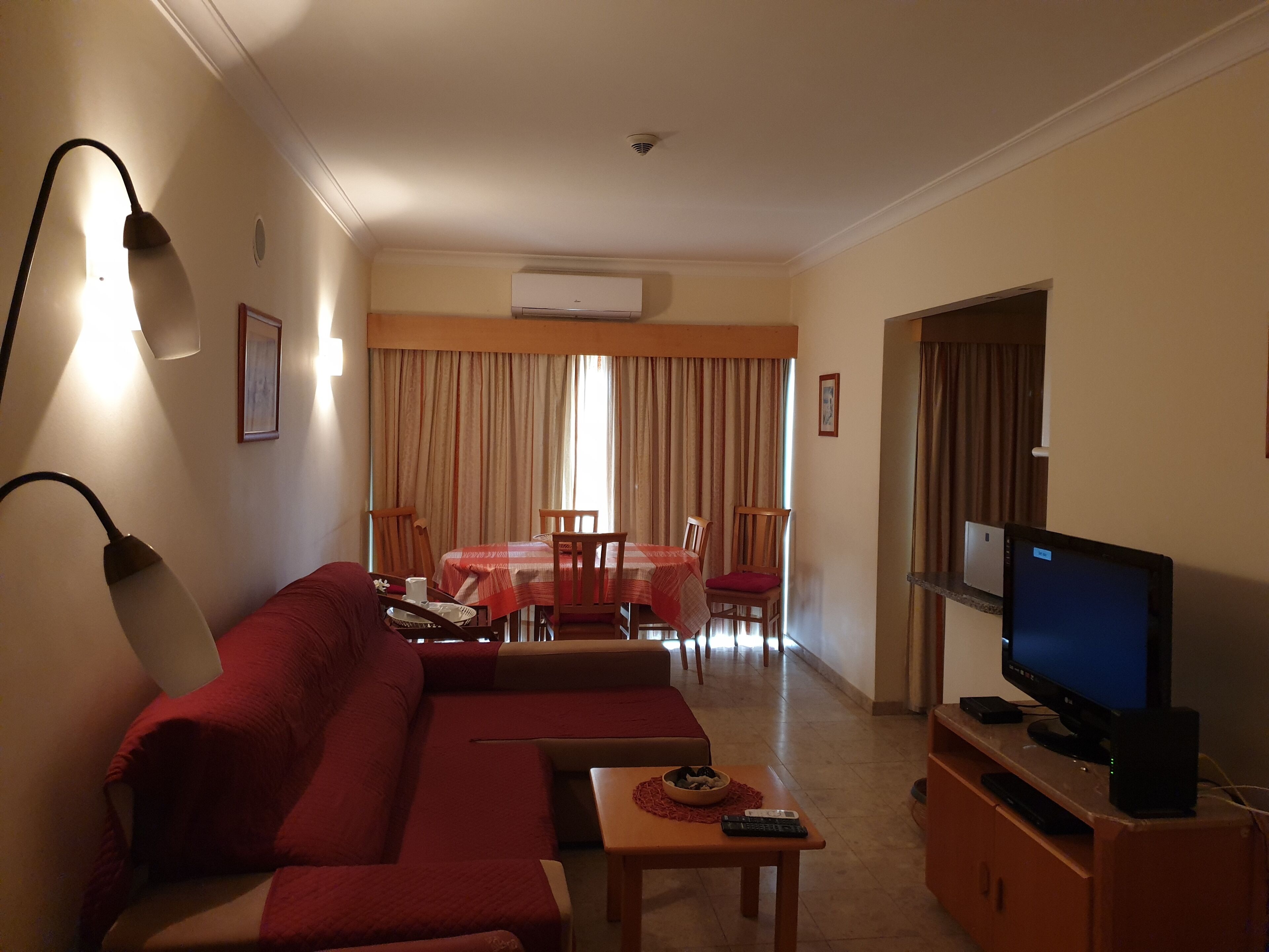 A living room with a red sofa, a TV on a wooden stand, a dining area with a table and chairs, and an air conditioning unit on the wall.