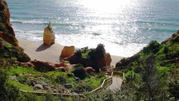 A coastal landscape with a sandy beach, rocky cliffs, and a wooden staircase leading down to the beach.