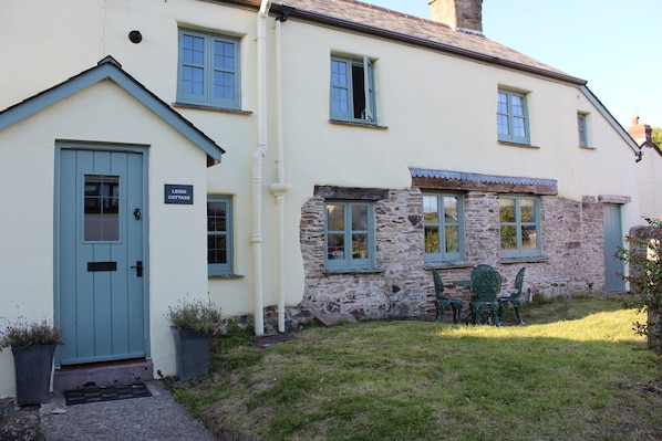 A two-story cottage with a blue door, stone wall, and a small garden with a table and chairs.