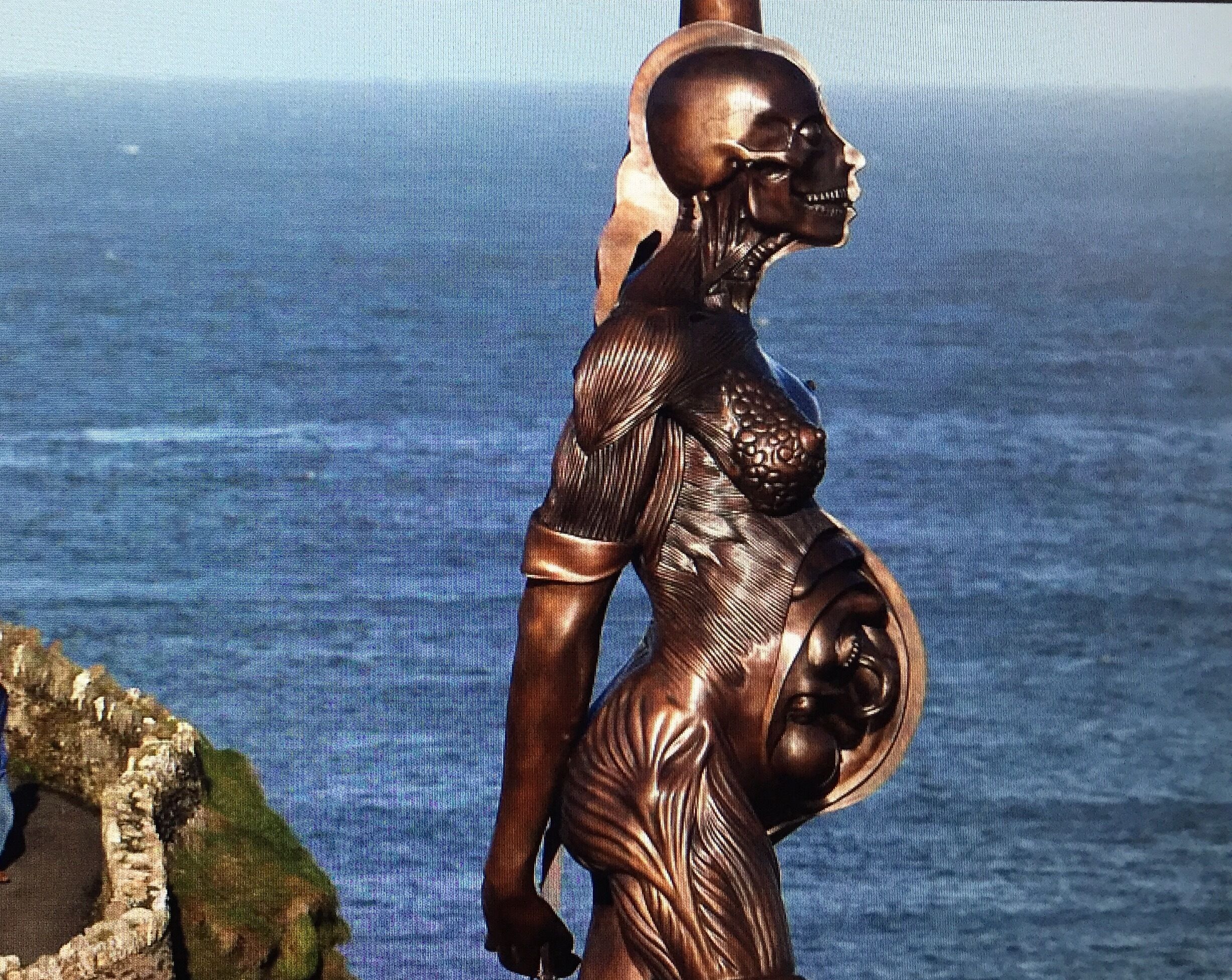 Escultura de una figura humana con rostro y cuerpo detallados, frente a una masa de agua y una costa rocosa.