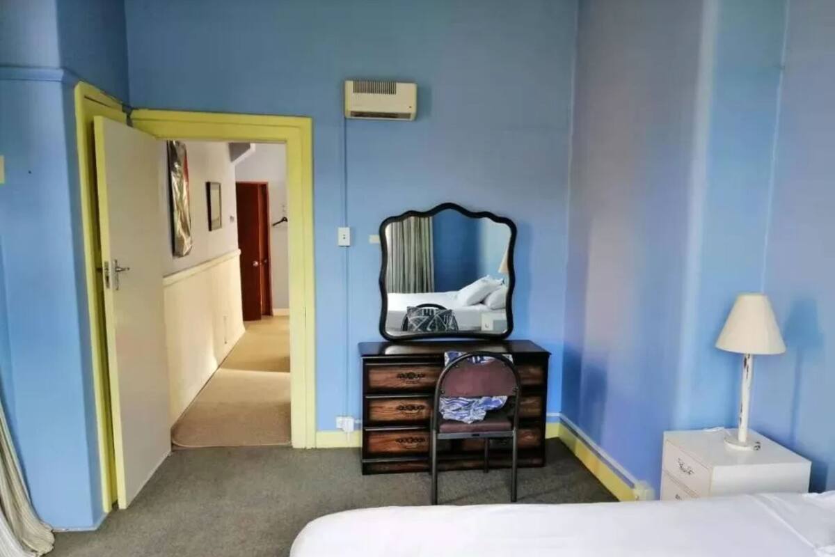 Una habitación de hotel con una cama, un aparador con espejo, una mesita de noche y una puerta que da a otra habitación.