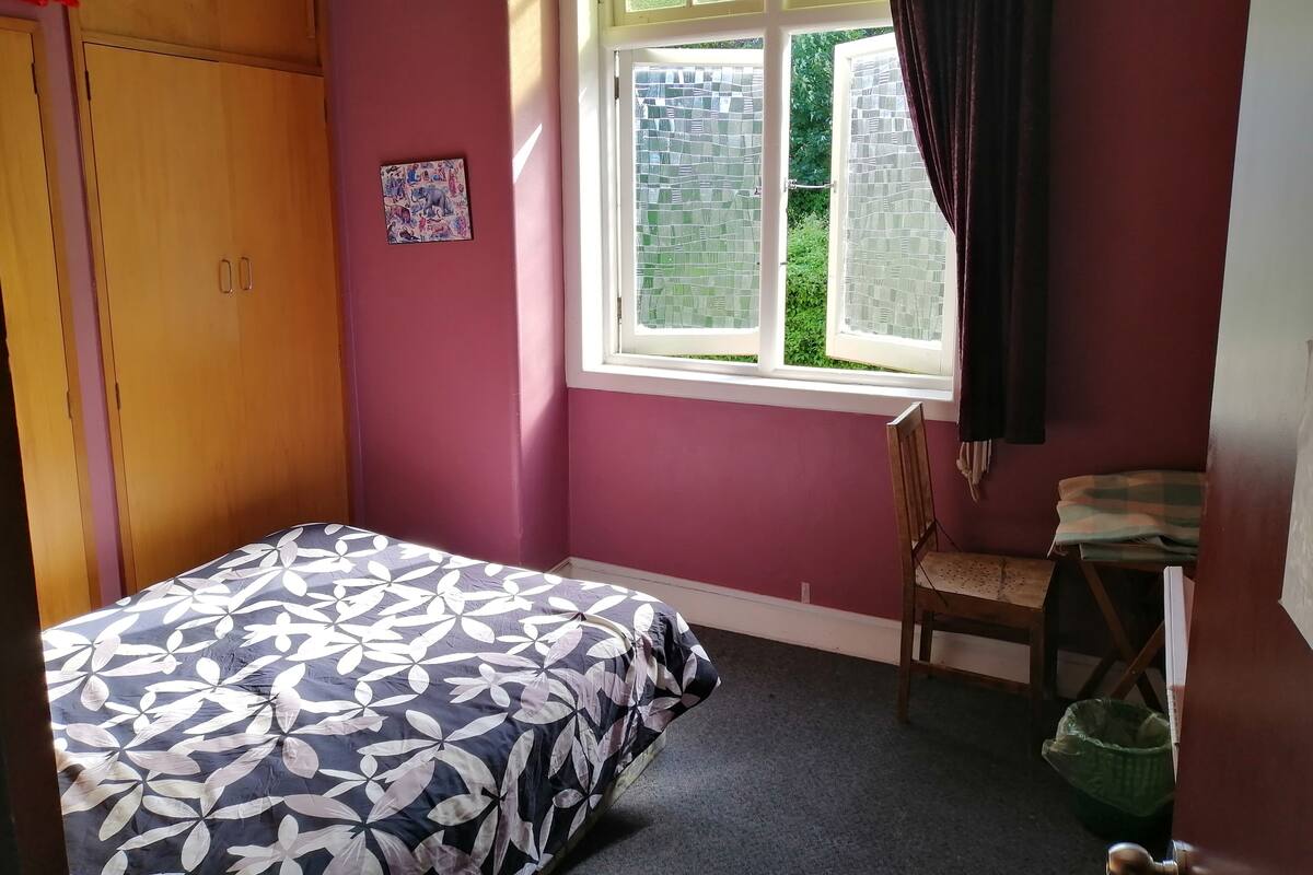 Un dormitorio con cama, ventana con cortinas, armario de madera y una silla.