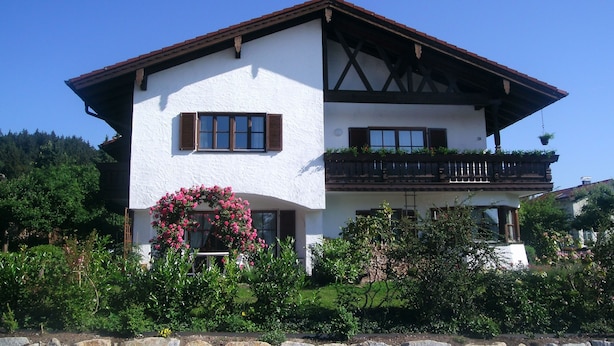 Une maison à deux étages, avec une façade blanche, des volets marron et un balcon agrémenté de plantes.