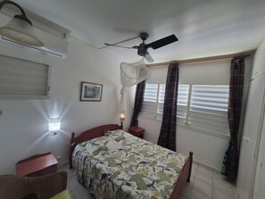 A bedroom with a bed, bedside tables, a ceiling fan, a wall-mounted air conditioner, a window with blinds, and a wall-mounted lamp.