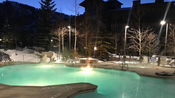 A swimming pool with a hot tub area, surrounded by a deck with chairs and a snow-covered landscape in the background.