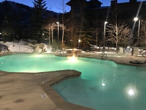 A swimming pool with a hot tub area, surrounded by a deck with chairs and a snow-covered landscape in the background.