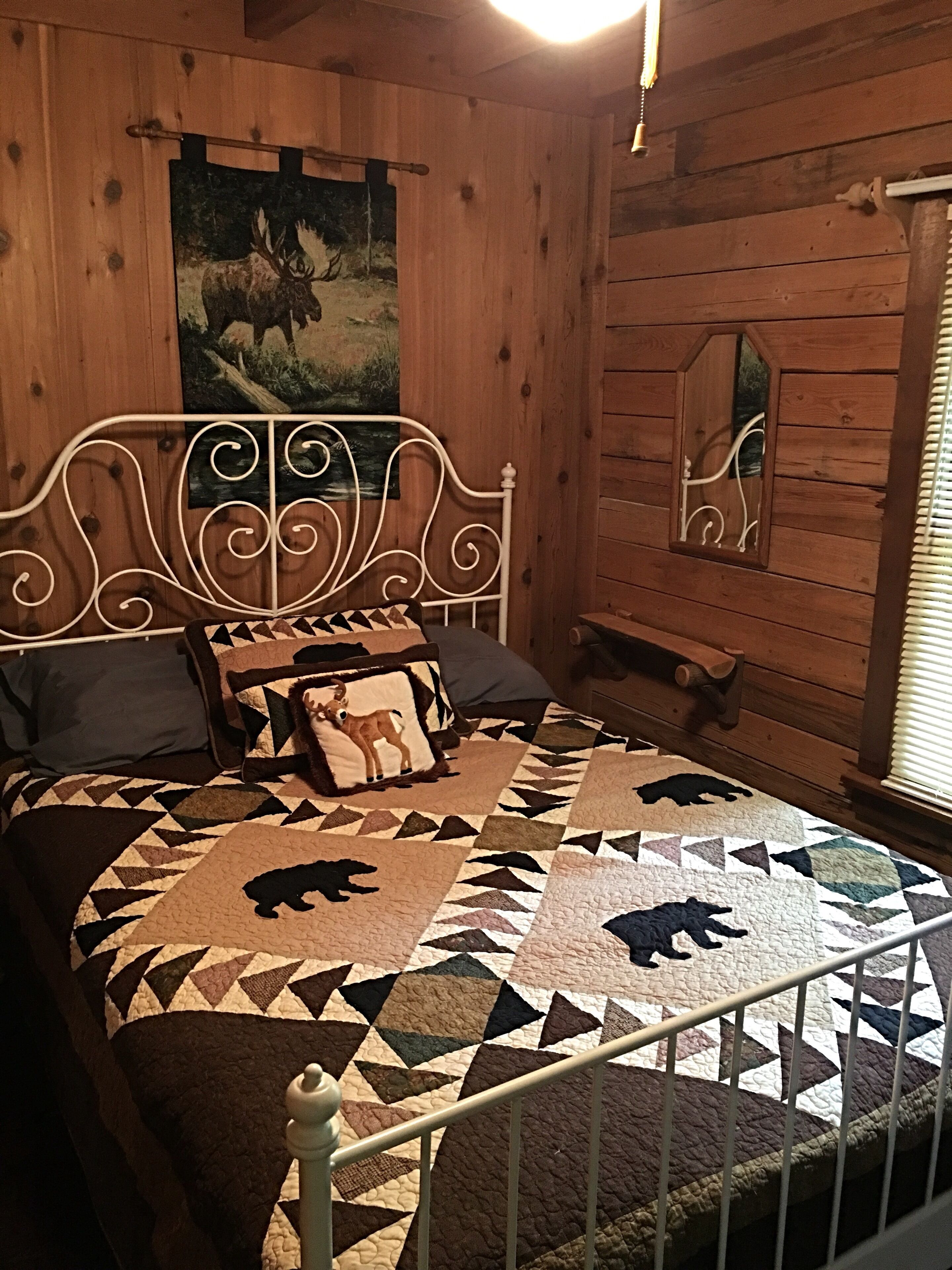 Ein Schlafzimmer einer Holzhütte mit einem Bett, einer Decke mit Tiermustern, einem Hirschbild und einem Fenster mit Jalousien.