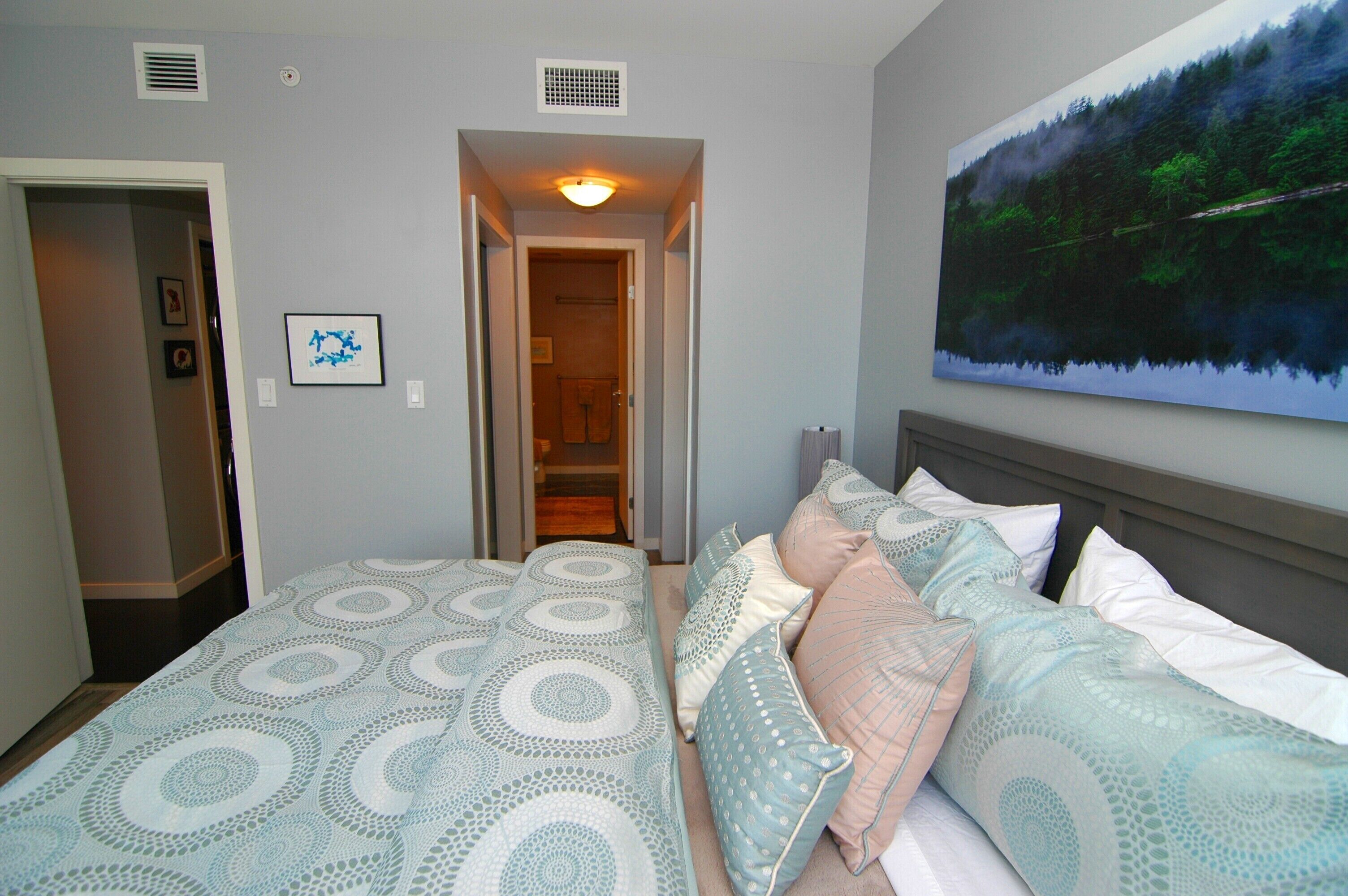 A bedroom with a bed, pillows, a painting, and a hallway leading to a bathroom.