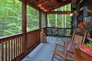 A wooden deck with a view of a forest, a table with a Coca-Cola box, and a chair with a striped pillow.