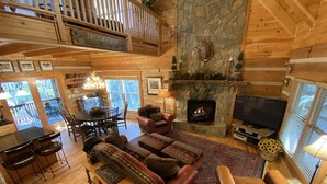 A cozy cabin interior with a stone fireplace, wooden furniture, and a balcony overlooking the outdoors.