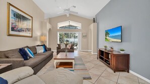 A spacious living room with a high ceiling, a ceiling fan, a large TV, and a view through an arched doorway to an outdoor area.