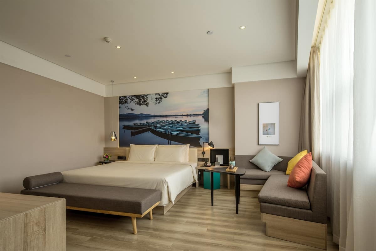 A modern hotel room with a large bed, a sofa, a small table, and a painting of boats on the wall.