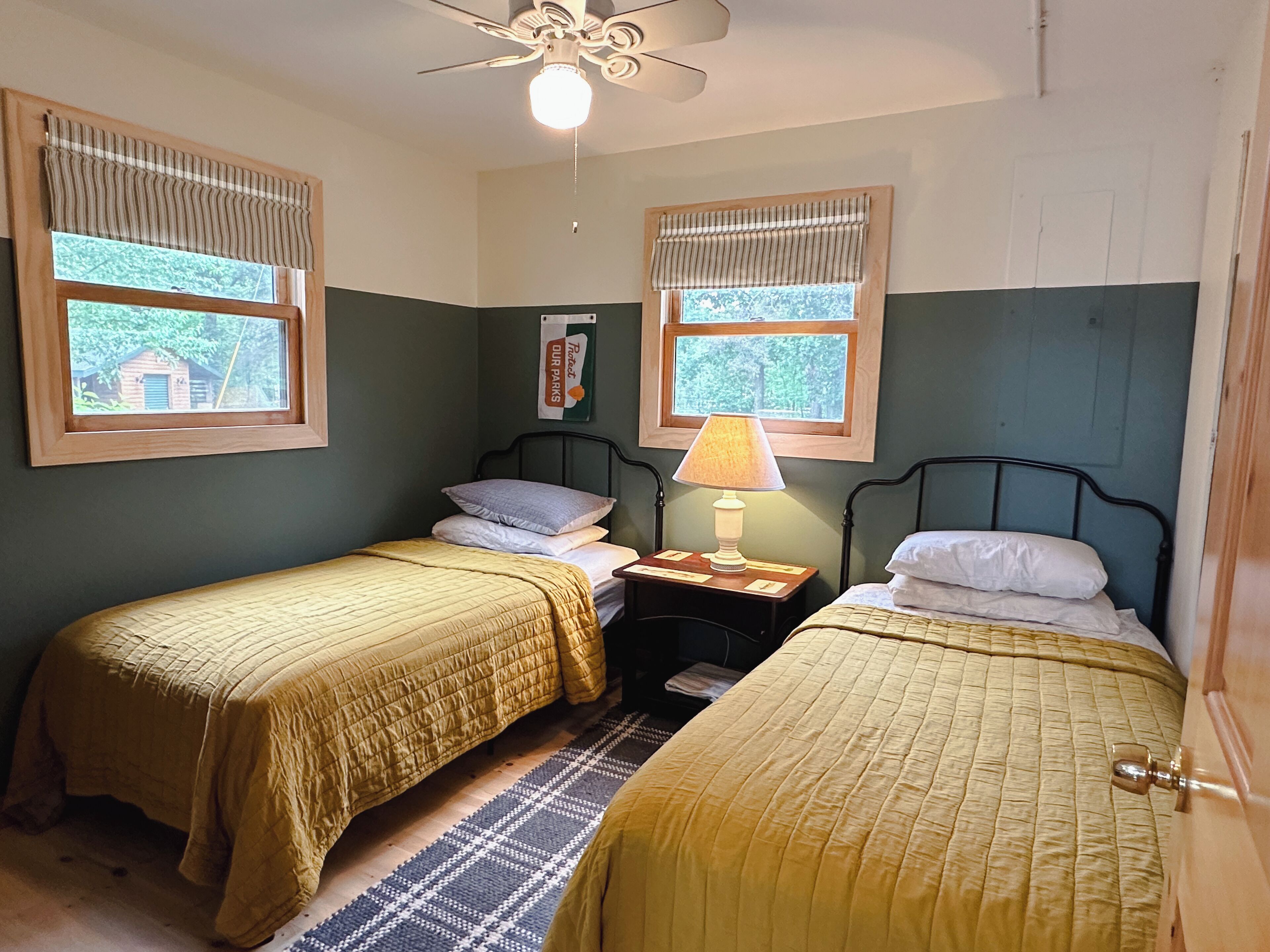 A room with two single beds, a ceiling fan, a window with a view of trees, and a sign on the wall.