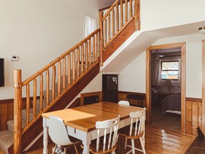 A dining area with a wooden table and chairs, a staircase with wooden railings, and a view of the outside through a window.