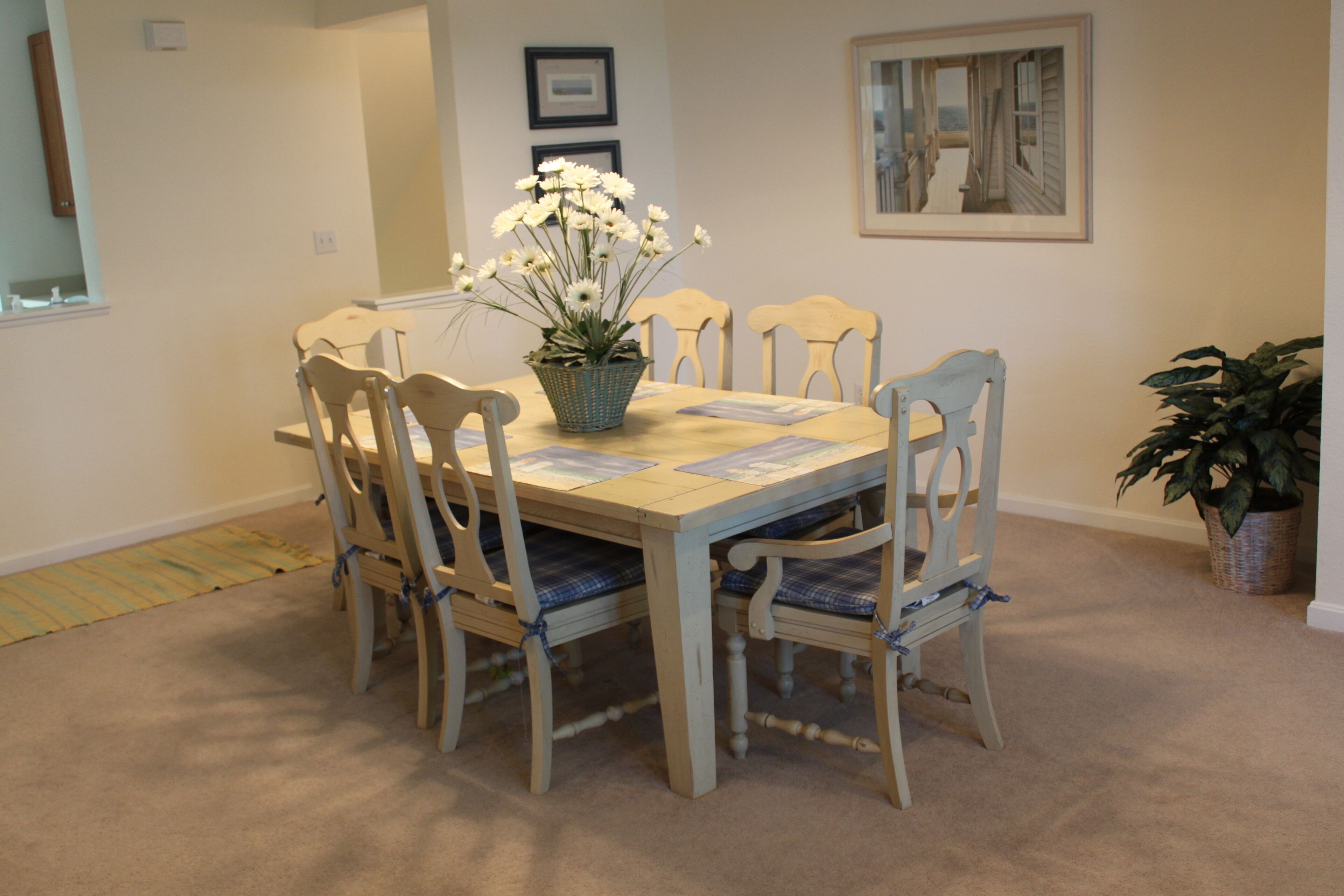 A dining area with a round table, six chairs, a vase of flowers, and a framed picture on the wall.