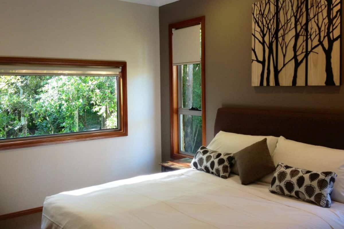 A bedroom with a large window, a bed with patterned pillows, and a wall-mounted artwork of trees.