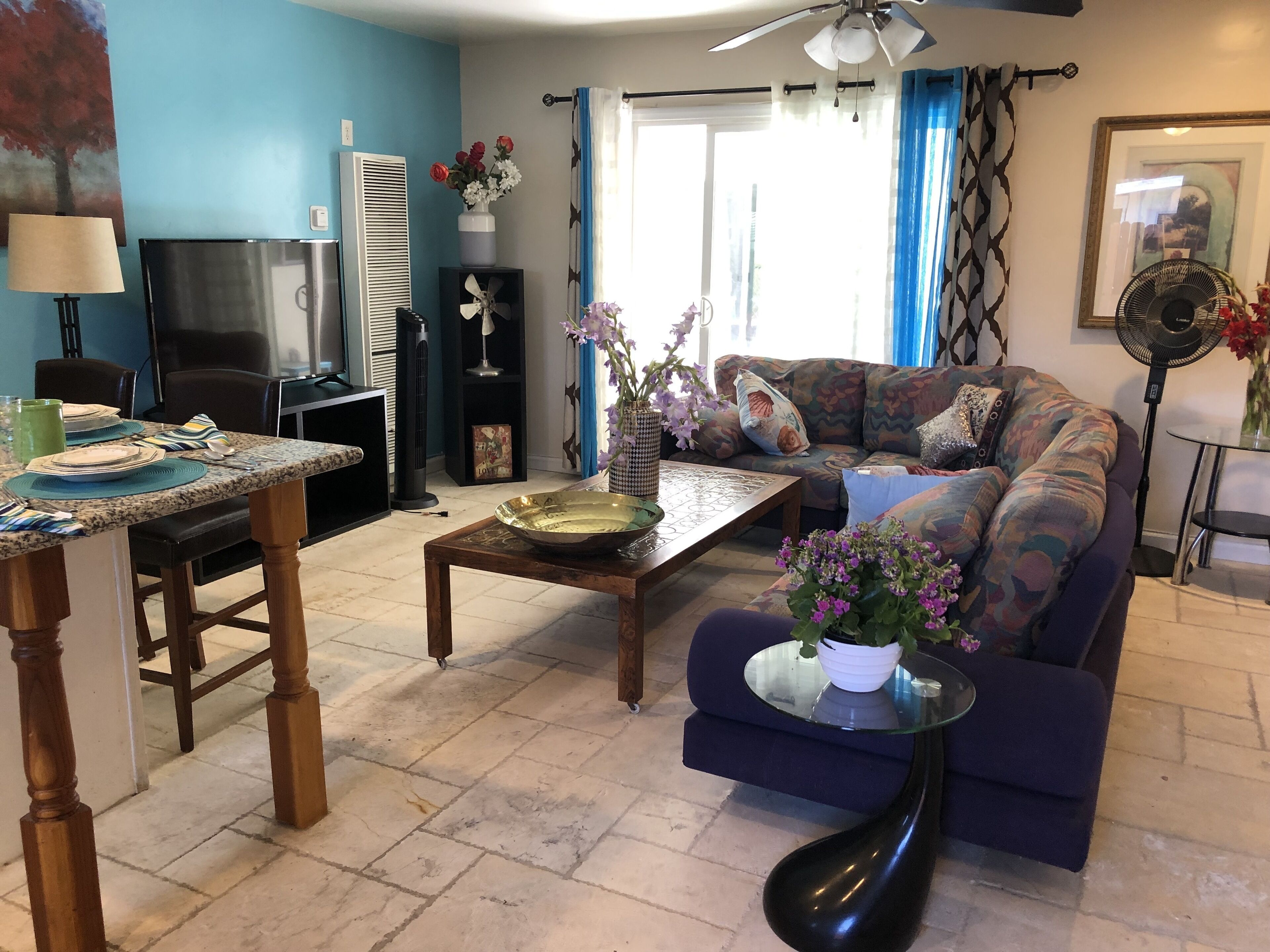 A living room with a sofa, a coffee table, a dining table with chairs, a TV, and a fan.