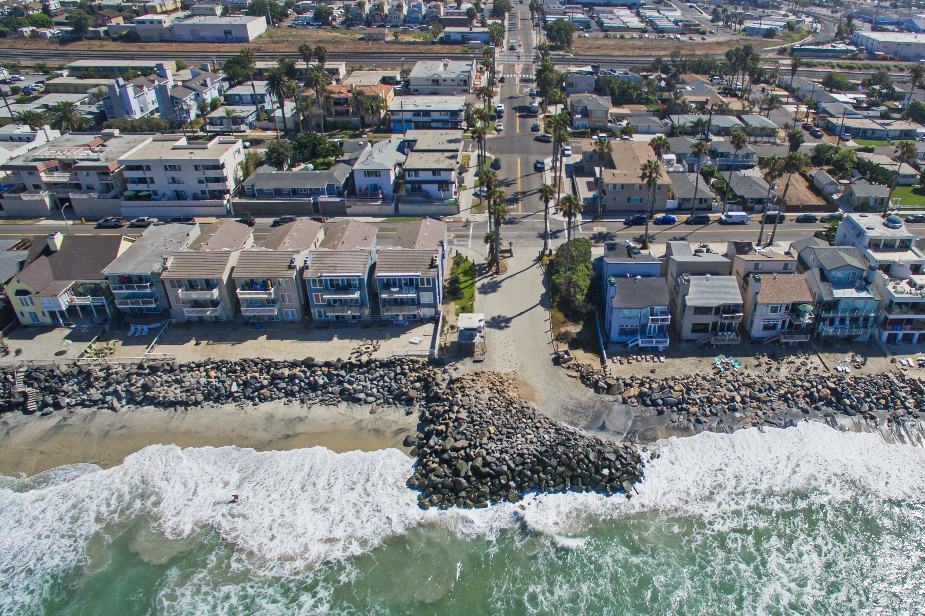 Vue aérienne d’un quartier résidentiel situé sur la côte, avec des propriétés donnant sur la plage, une digue rocheuse et une route dégagée menant à la plage.