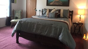 Quarto com uma cama grande, porta de madeira, cadeira, quadro, luminária e uma pequena mesa.