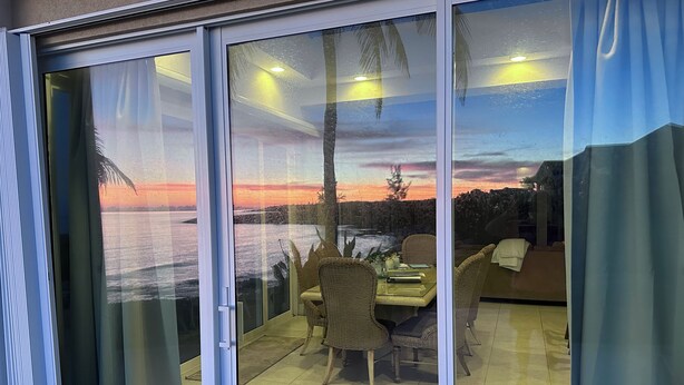 A room with a sliding glass door leading to a patio area with wicker furniture and a view of a sunset over water. - Secluded Waterfront Single Family Villa with Private Pool – 3,000 Sq of Luxury (East End - Half Moon Bay)