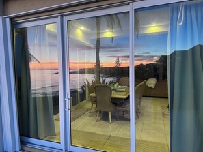 A room with a sliding glass door leading to a patio area with wicker furniture and a view of a sunset over water.