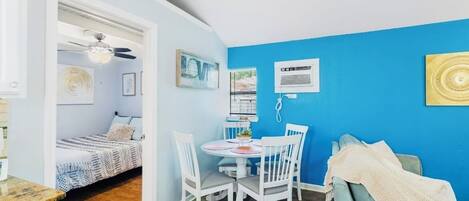 A room with a bed, a dining table with chairs, a ceiling fan, and a blue wall with framed pictures.