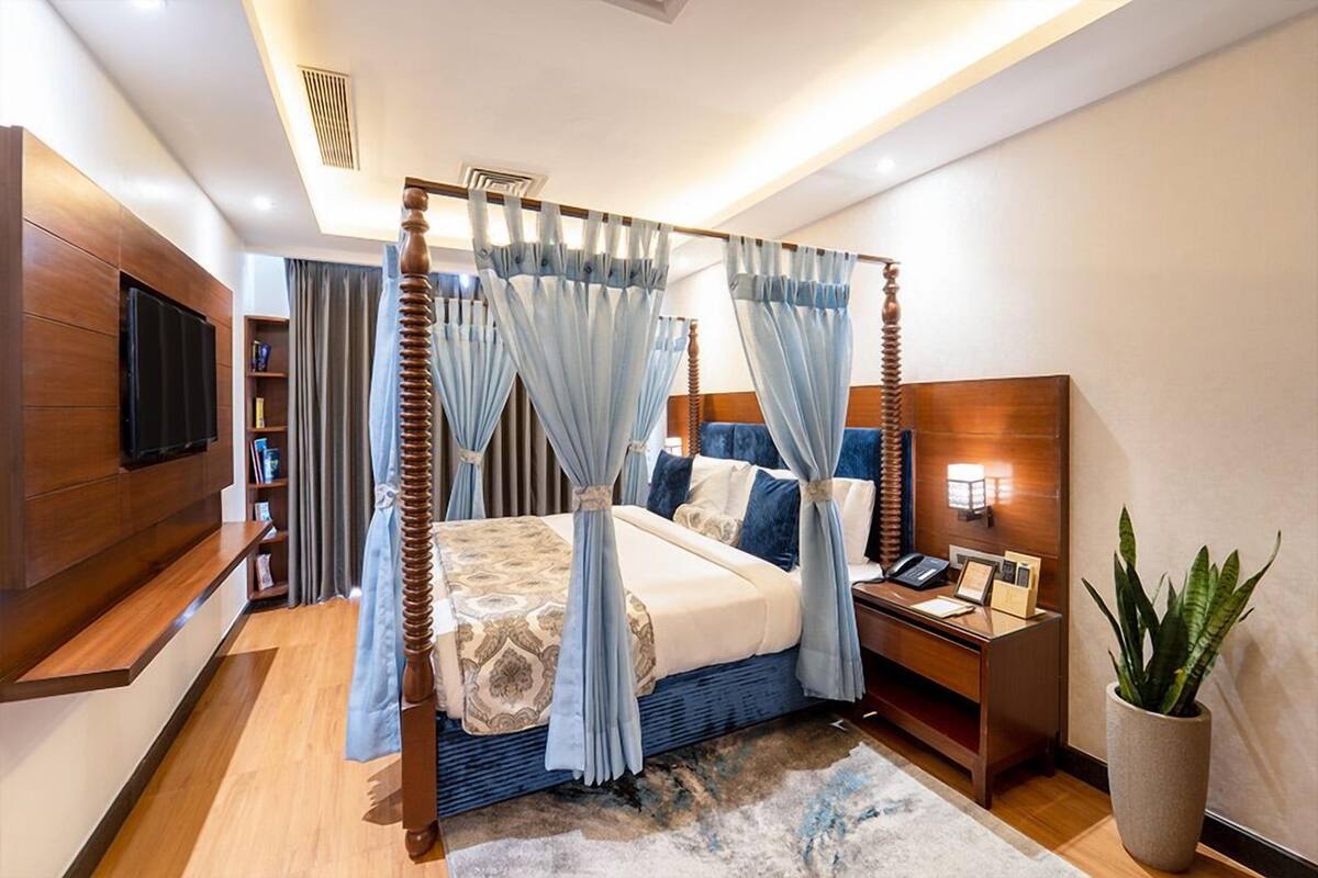 Quarto moderno com cama de dossel, mesa de cabeceira com abajur, televisão na parede e planta em vaso.