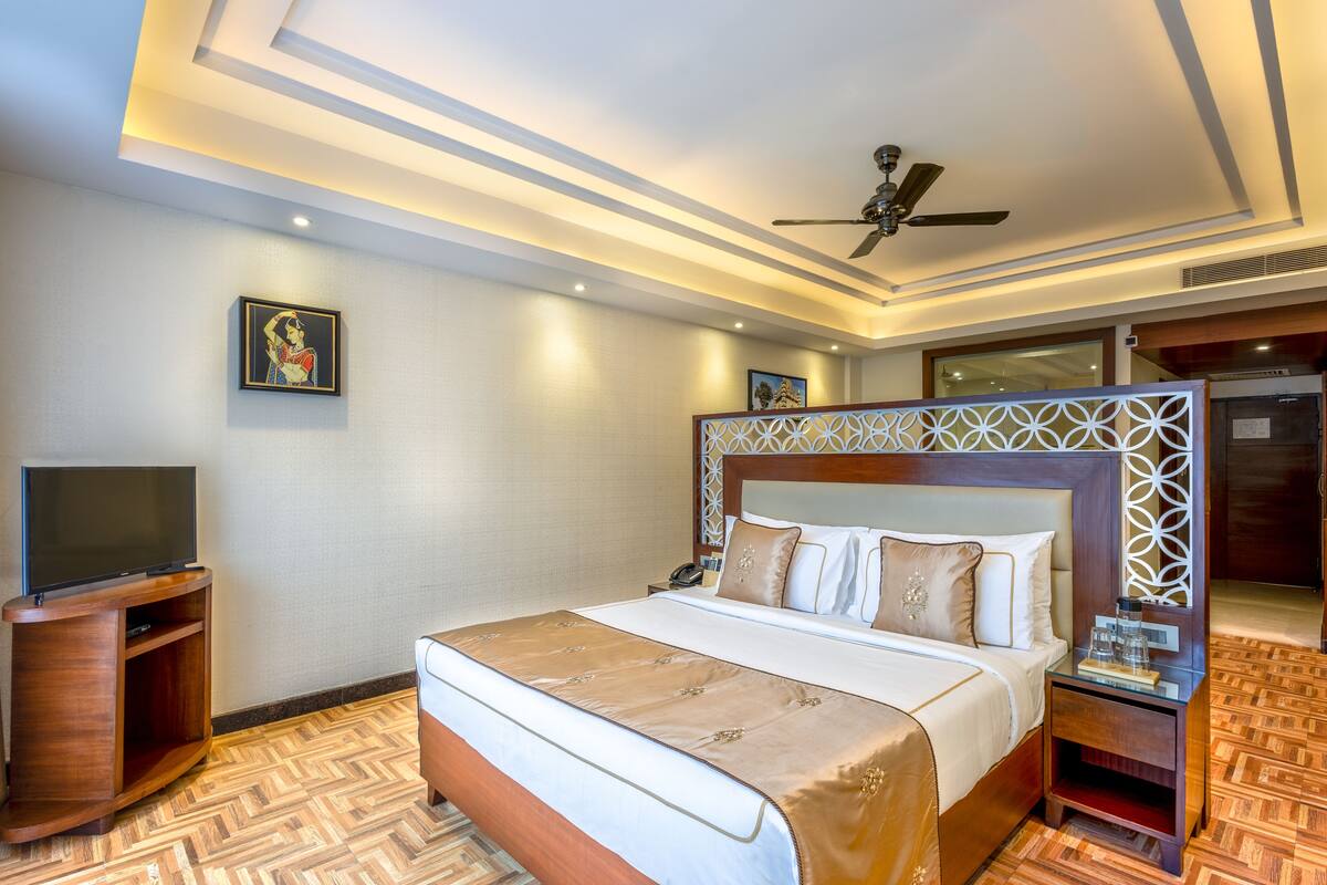 Quarto de hotel com uma cama grande, televisão, mesa de cabeceira de madeira e ventilador de teto.