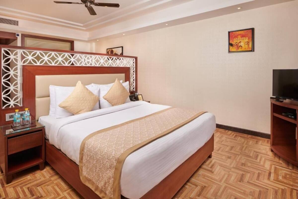 Quarto de hotel com cama grande, televisão, piso de madeira e cabeceira decorativa.