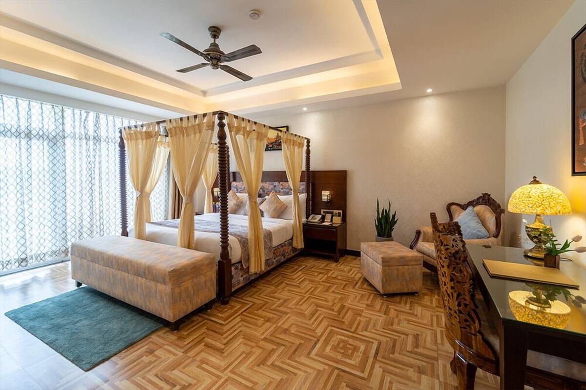 Uma cama de dossel com cortina, piso de madeira, ventilador de teto, mesa de cabeceira com abajur e vista para o exterior por grandes janelas.