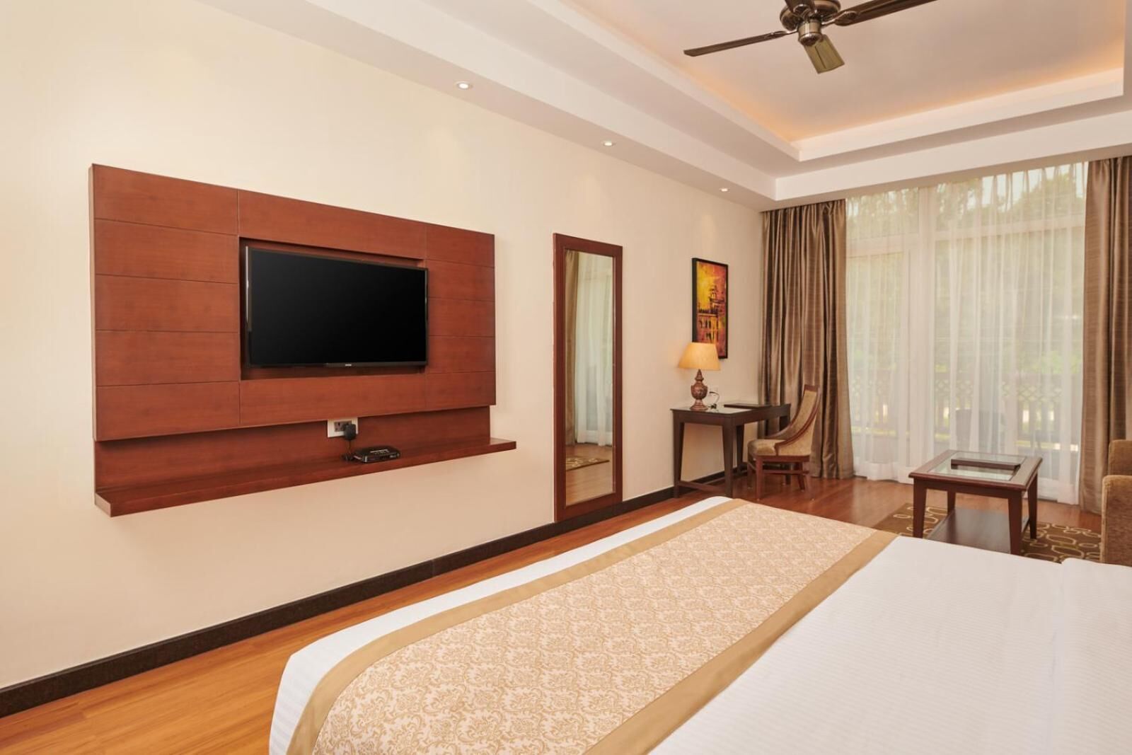 A hotel room with a large bed, a mounted TV, a desk with a chair, a lamp, and a window with curtains.