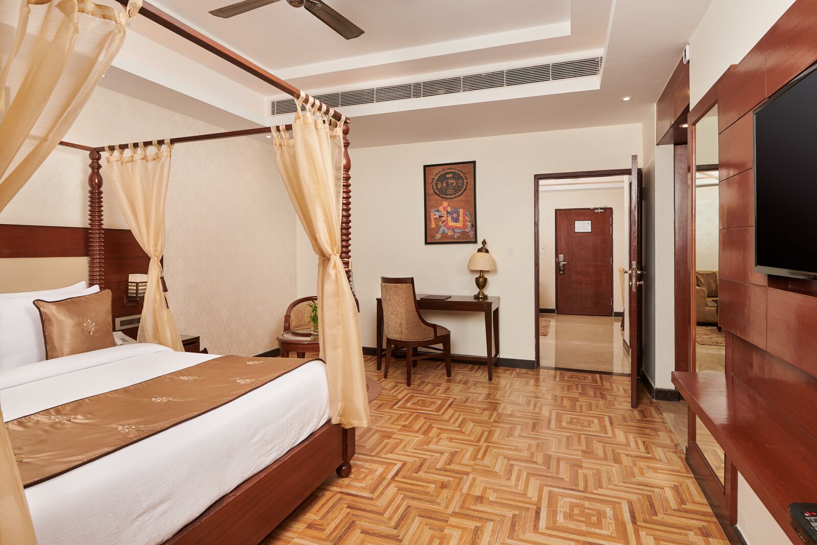 A hotel room with a four-poster bed, a desk with a chair, a television, and a door leading to another room.