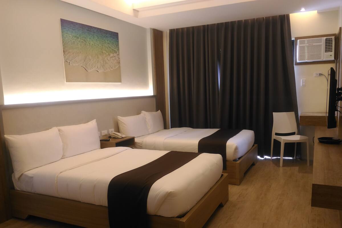 A hotel room with two beds, a painting on the wall, a curtain, a desk with a chair, and an air conditioning unit.