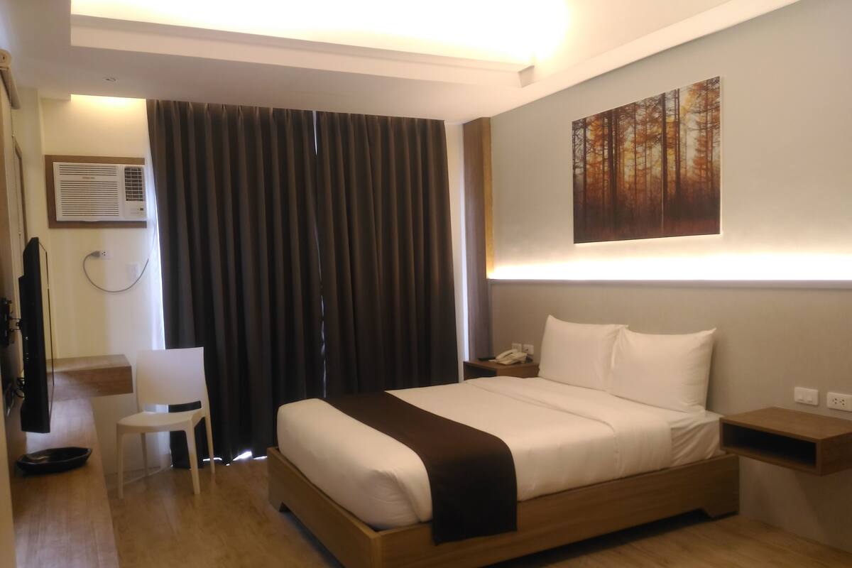 A hotel room with a bed, bedside table, chair, and a painting on the wall.
