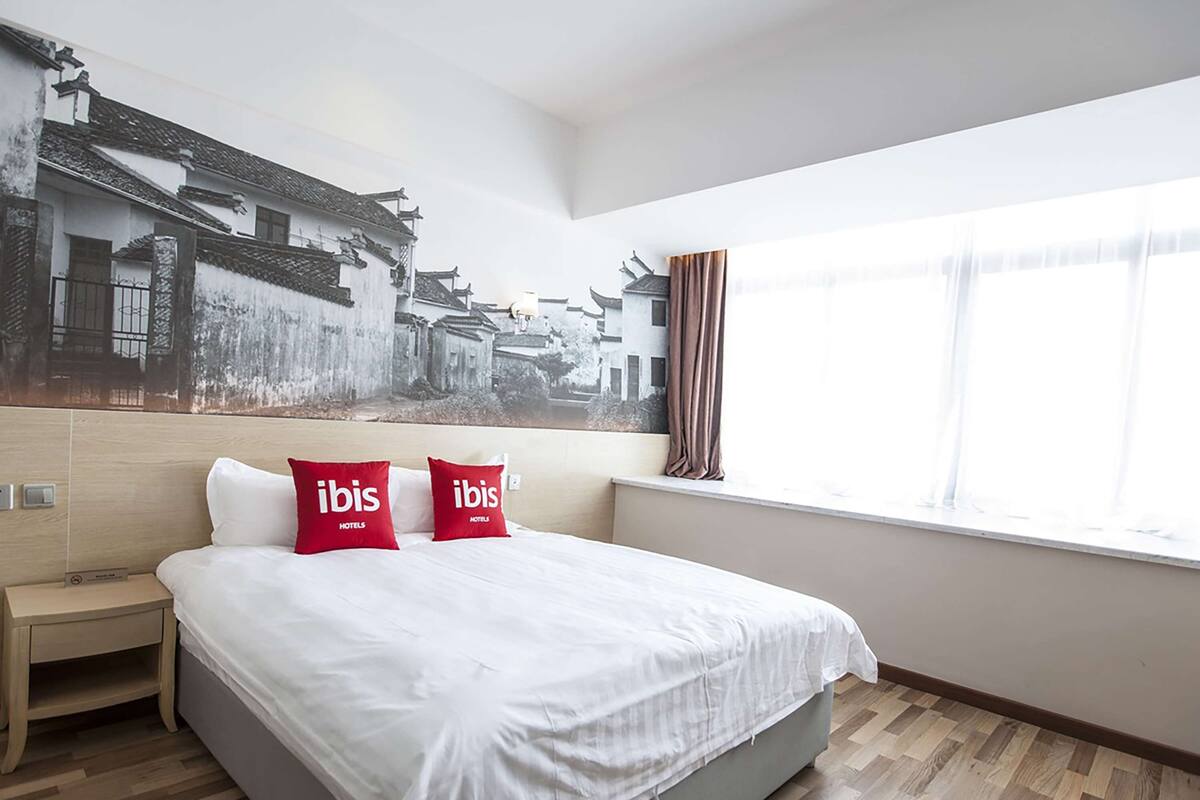 A hotel room with a large bed, two red pillows with "ibis Hotels" branding, a wooden nightstand, a large window with curtains, and a wall mural of traditional buildings.