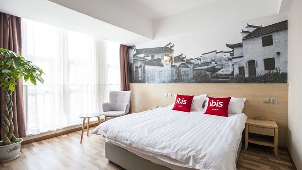 A hotel room with a large bed, a chair, a nightstand, and a wall-mounted painting of traditional buildings. - ibis Kunshan Changjiang Rd Hotel (Suzhou)