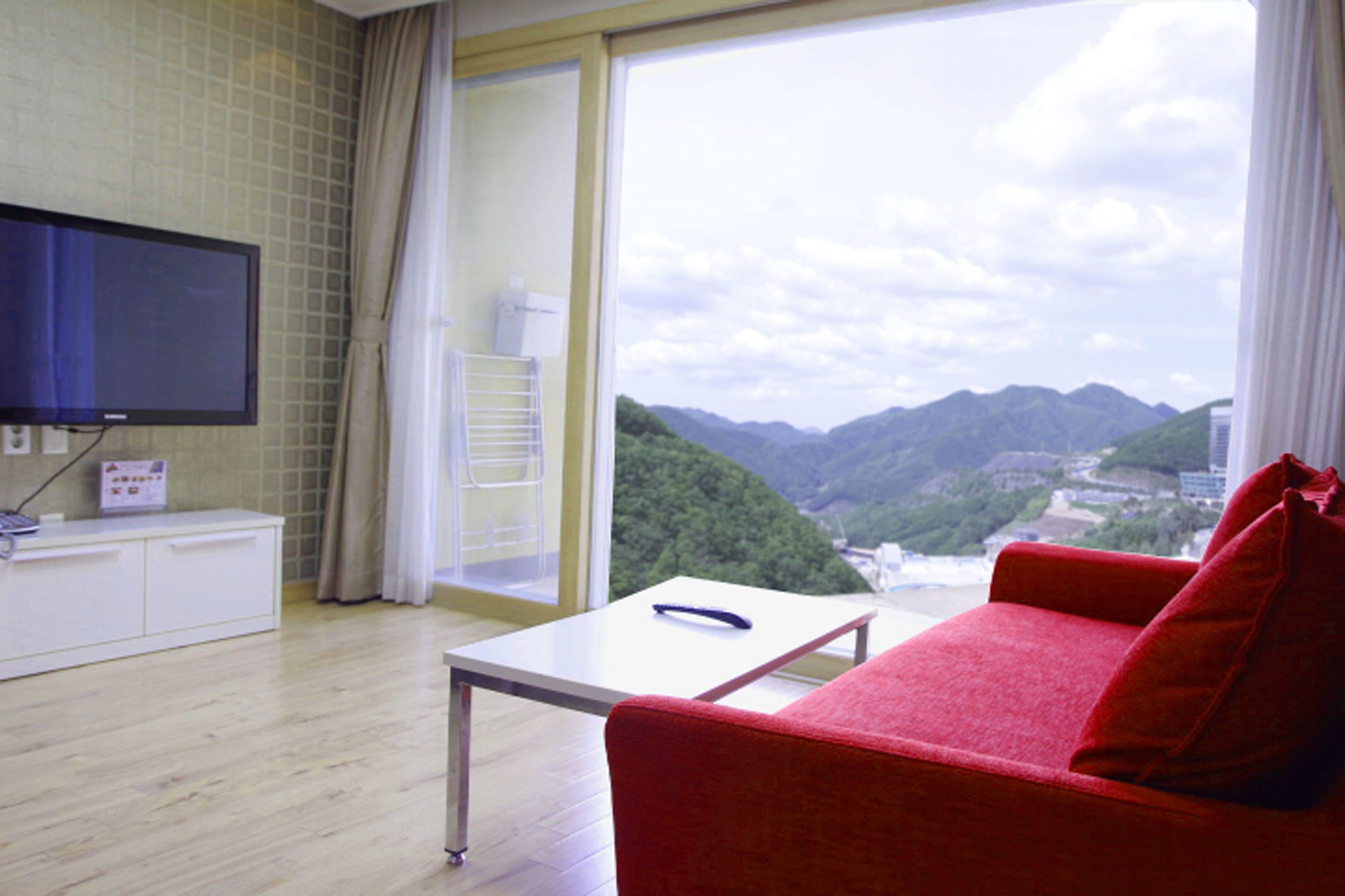 A hotel room with a red sofa, a TV mounted on the wall, a table, and a view of mountains through the window.