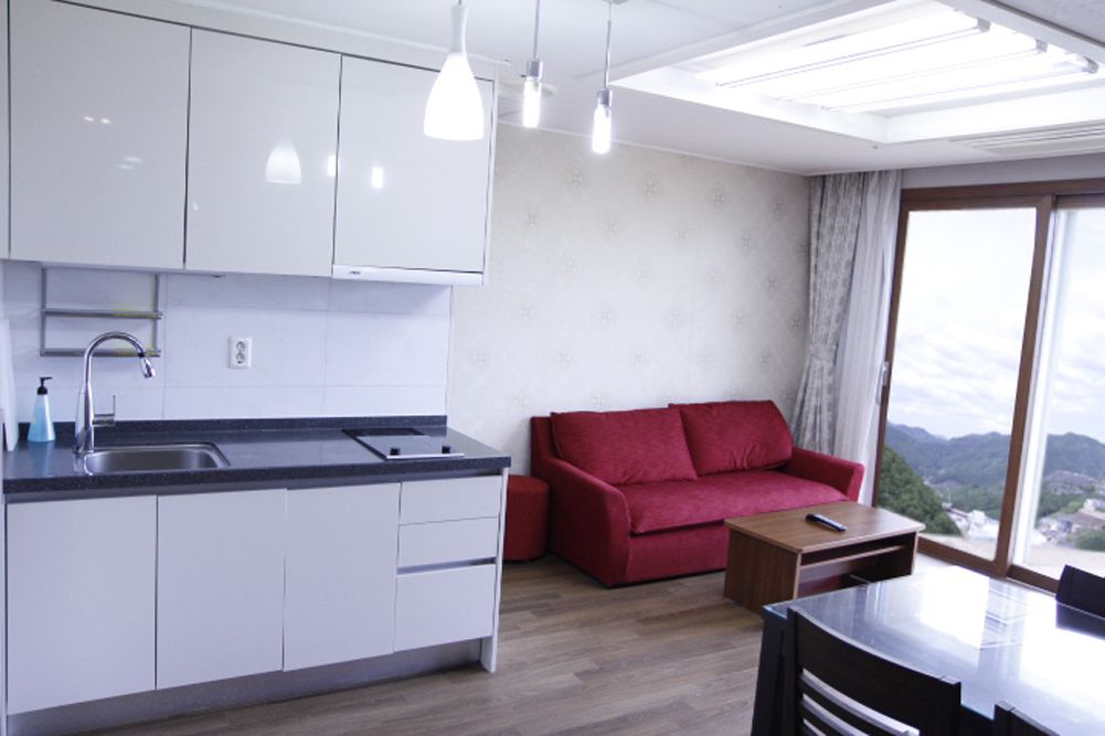 Una cocina moderna con armarios blancos, una encimera negra y un fregadero. Hay un sofá rojo y una pequeña mesa de madera con un control remoto. Un amplio ventanal ofrece vistas al exterior.