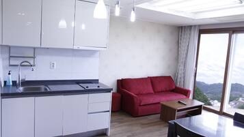 Una cocina moderna con armarios blancos, una encimera negra y un fregadero. Hay un sofá rojo y una pequeña mesa de madera con un control remoto. Un amplio ventanal ofrece vistas al exterior.