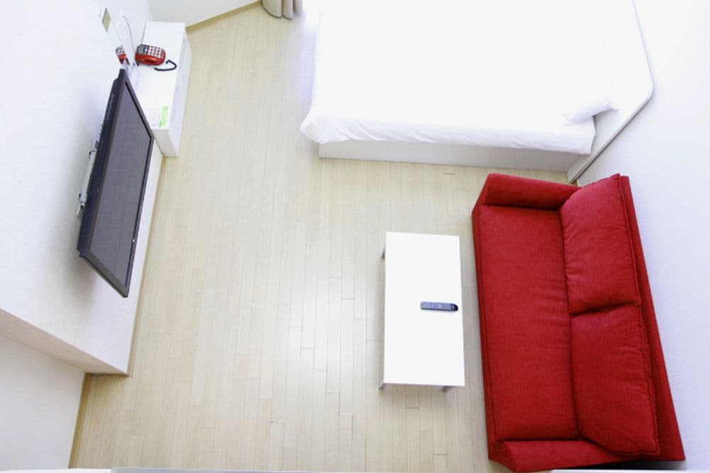 A room with a bed, a red sofa, a white bedside table, and a flat-screen TV mounted on the wall.