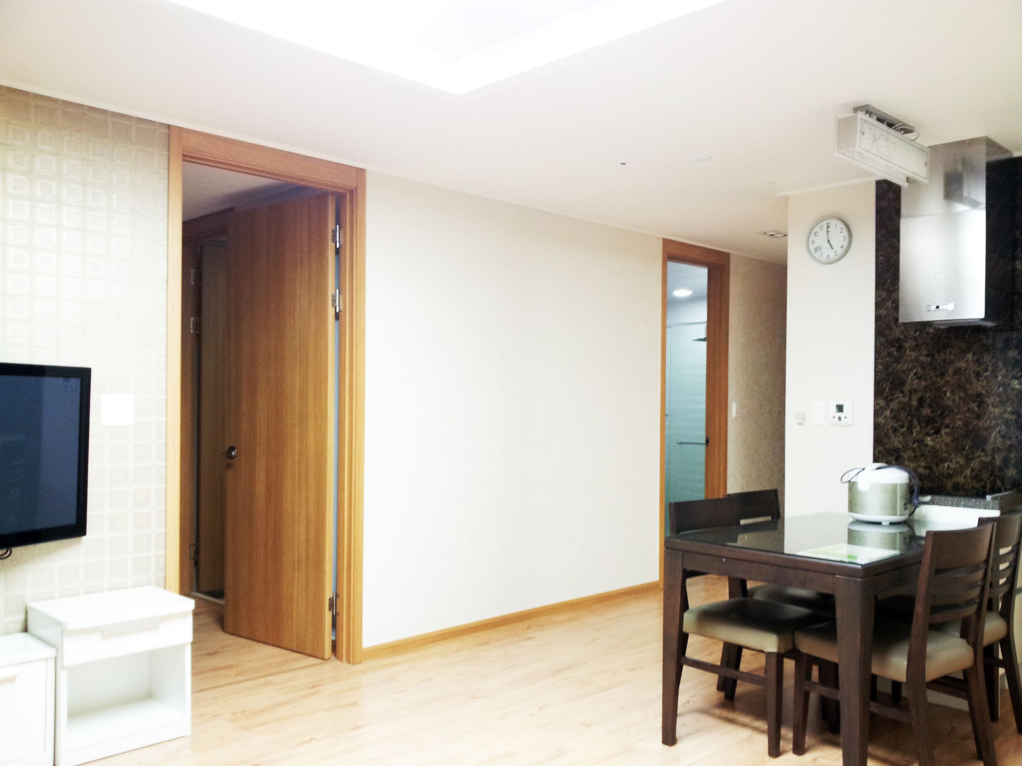A dining area with a table and chairs, a television mounted on the wall, and a kitchen area with a clock and an air conditioning unit.
