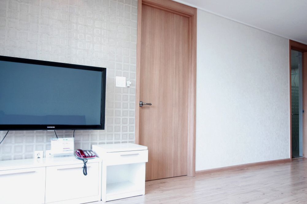 A modern room with a flat-screen TV mounted on a white cabinet, a wooden door, and a light-colored tiled wall.