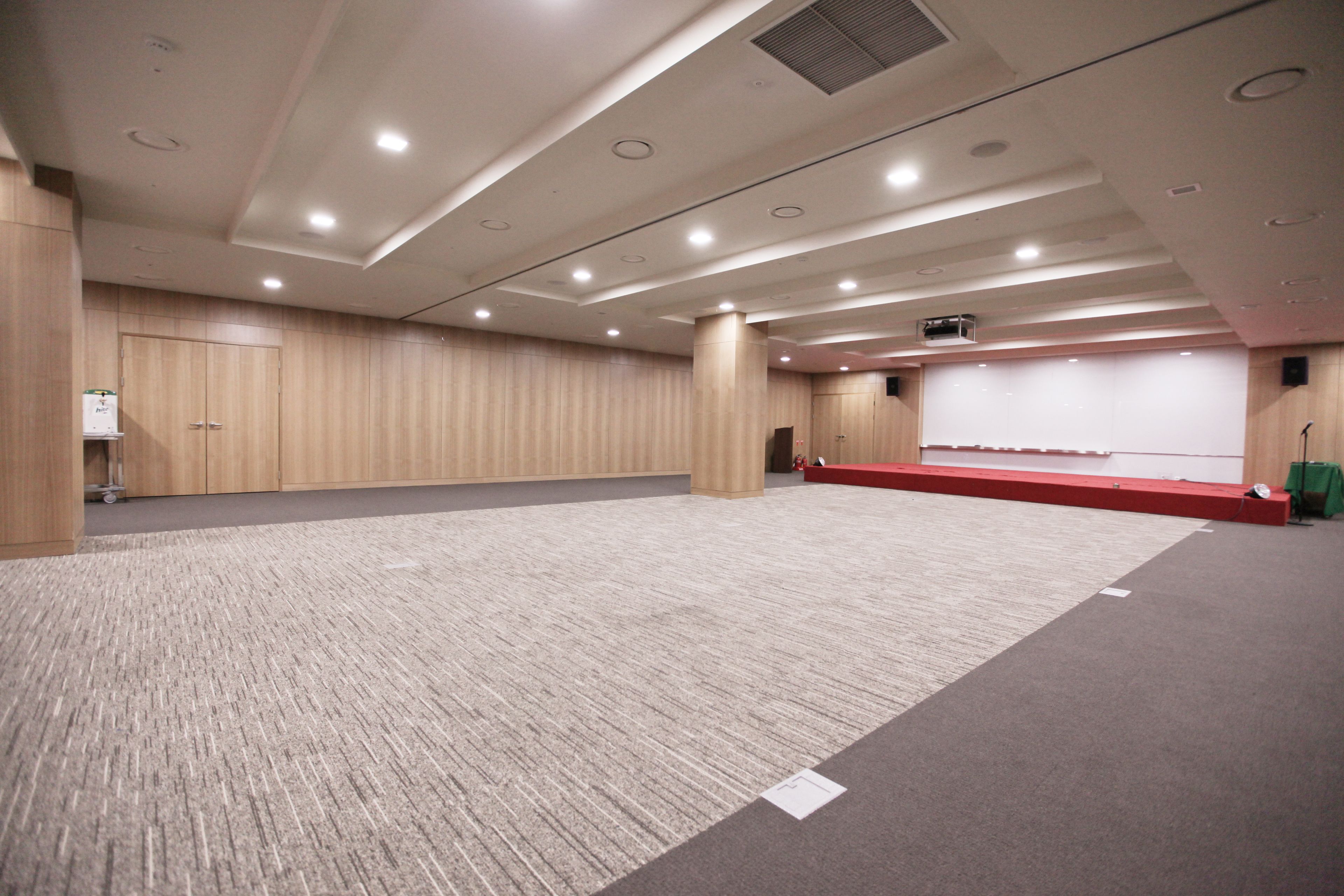 A conference room with a carpeted floor, wooden panel walls, a stage with a red carpet, and ceiling lights.