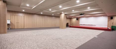 A conference room with a carpeted floor, wooden panel walls, a stage with a red carpet, and ceiling lights.