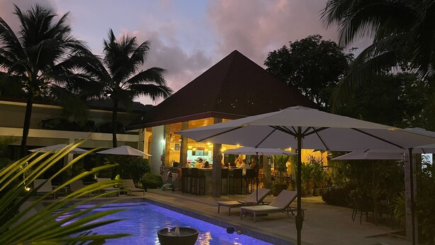A pool area with lounge chairs and umbrellas, a bar, and a building with a red roof. - Lotus Bleu Resort & Restaurant (Rawai)