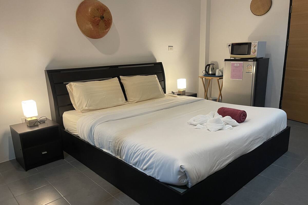 A neatly made bed with white linens, two bedside tables with lamps, a microwave, and a small table with a kettle.